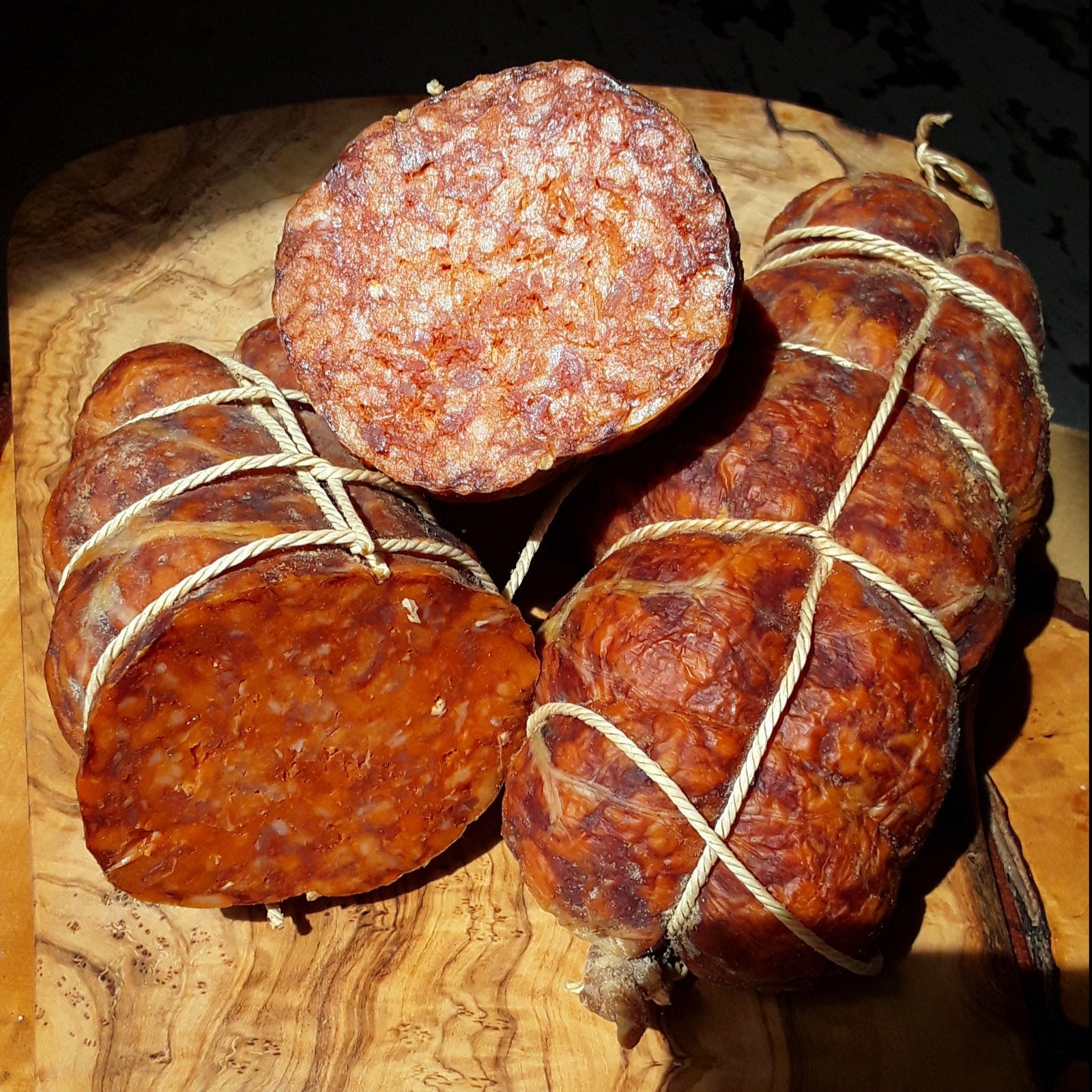 Several pieces of Mangalitsa Nduja salami are displayed on a wooden board, tied with strings, with visible spicy and richly colored meat mixture.